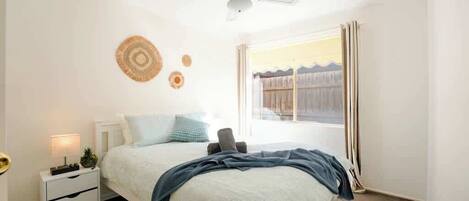 Un dormitorio con cama, mesita de noche, ventilador de techo y ventana con cortinas.
