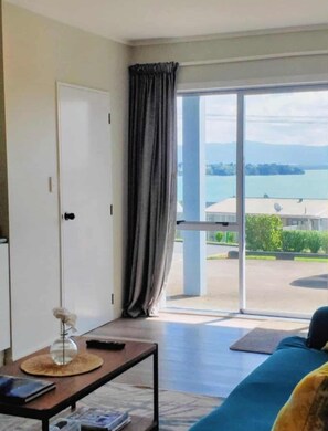 A room with a large window, a sofa, a table with a lamp, and a view of a body of water and buildings. - Modern family friendly apartment with great views, few steps away to the sea (Ōmokoroa)