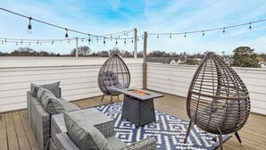 Une terrasse sur le toit, aménagée avec des meubles en osier, une table dotée d’un foyer intégré et illuminée de guirlandes lumineuses.