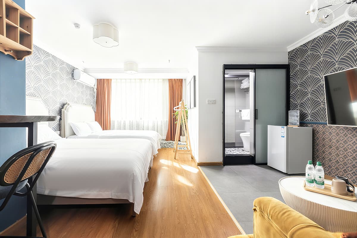 A hotel room with two beds, a desk, a chair, a TV, and a small table with water bottles.