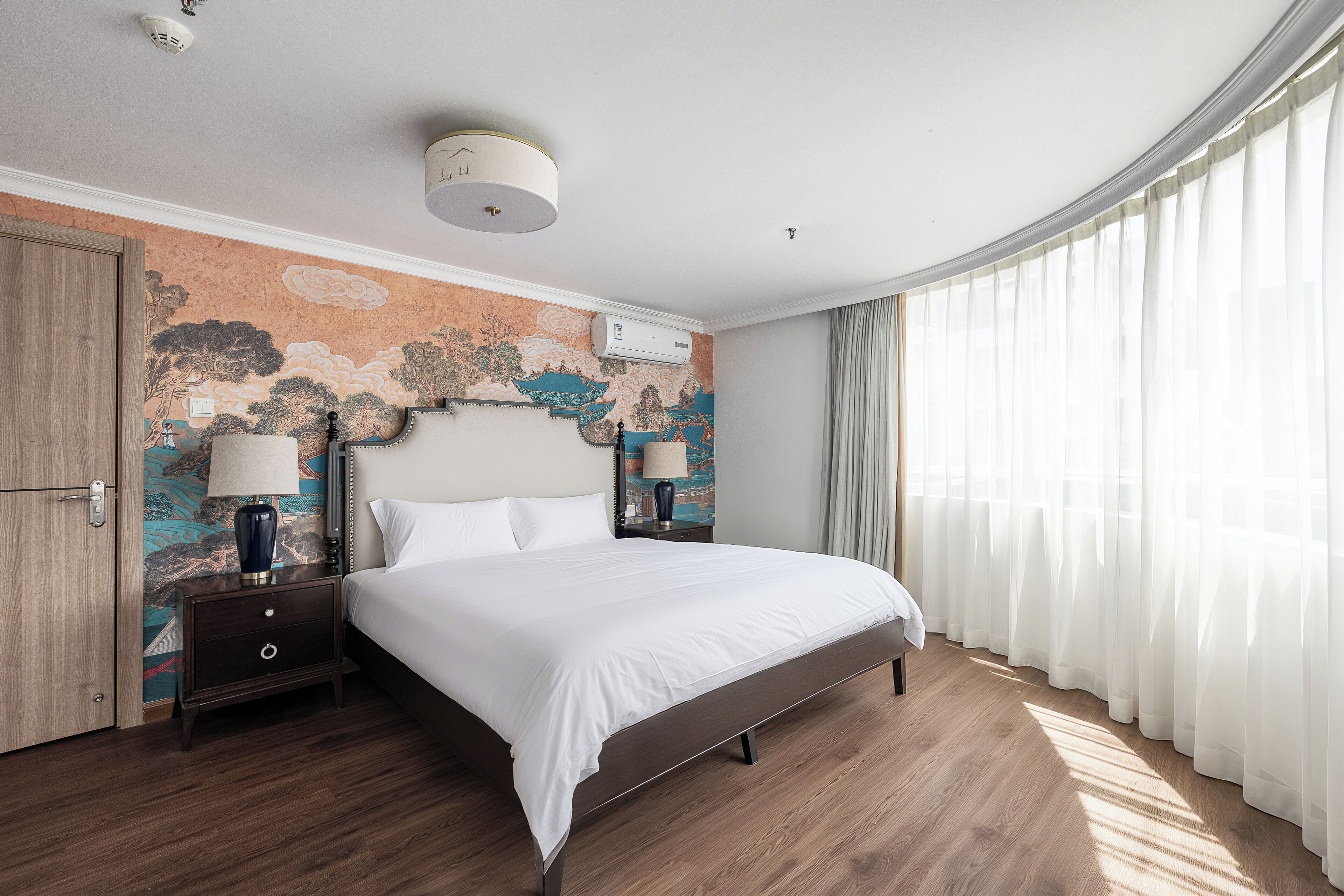 A hotel room with a large bed, a nightstand with a lamp, a wall mural, and a window with sheer curtains.