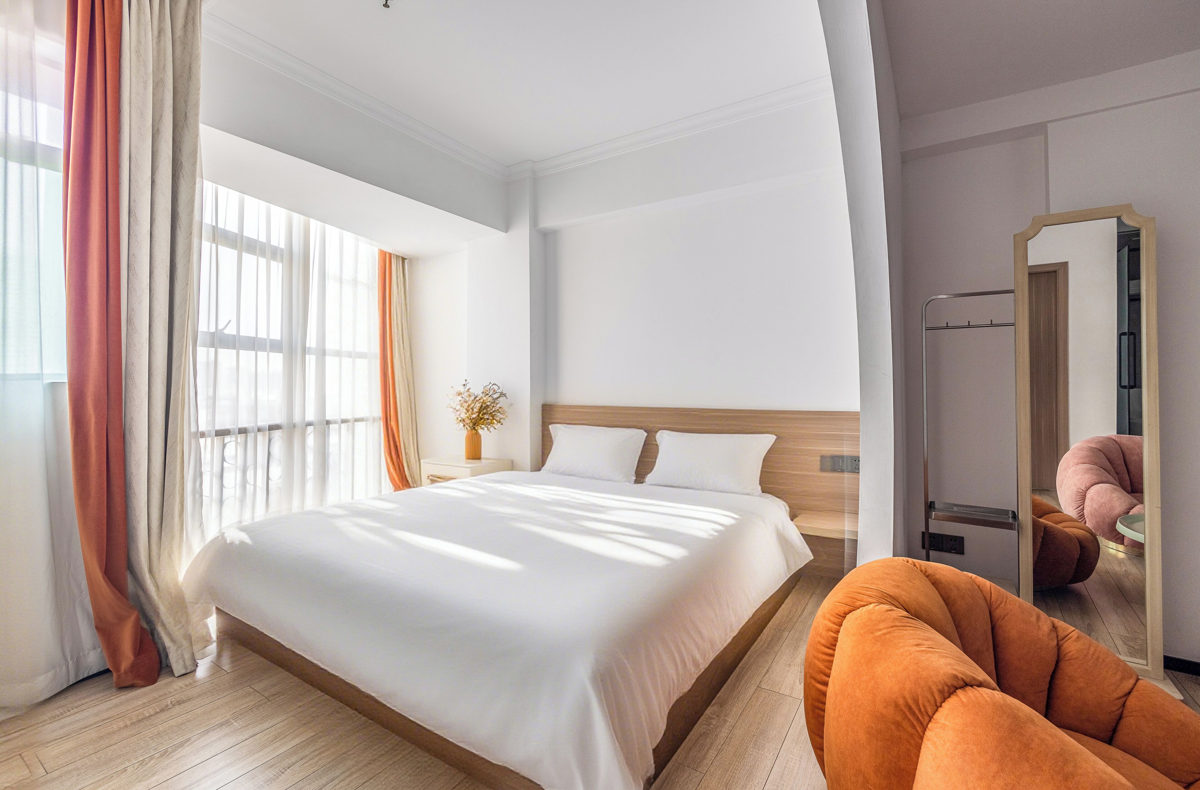 A modern hotel room with a large bed, a wooden headboard, a mirror, and a seating area with a chair.