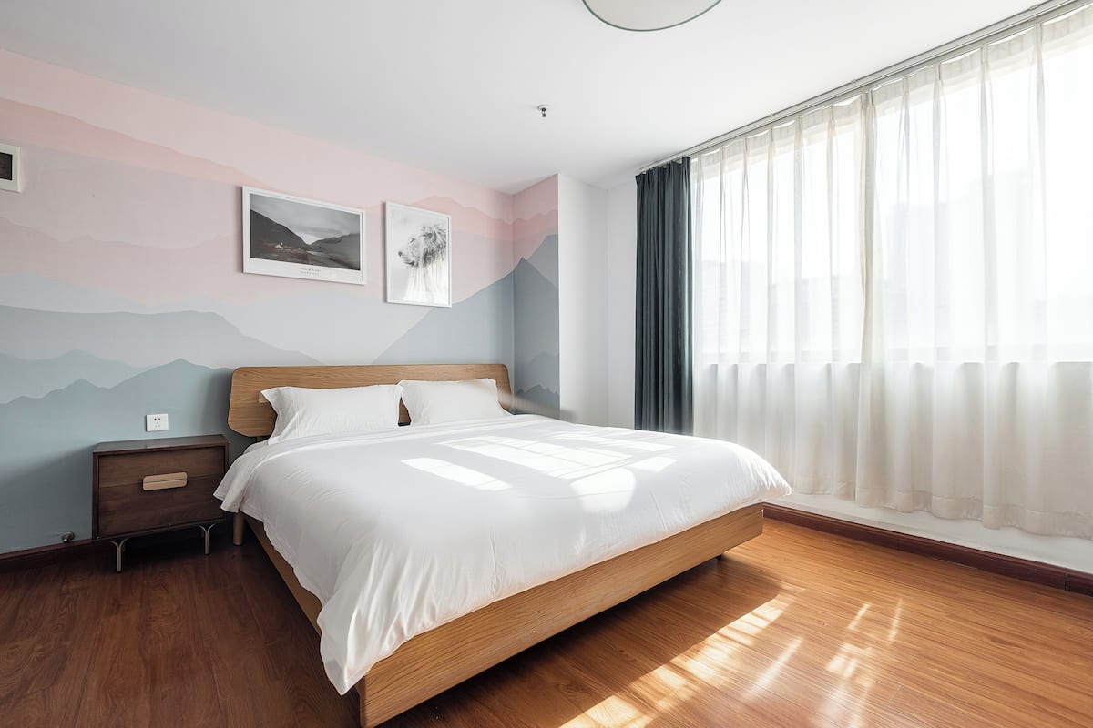 A bedroom with a wooden bed, a nightstand, a wall with a mountain design, and a window with sheer curtains.