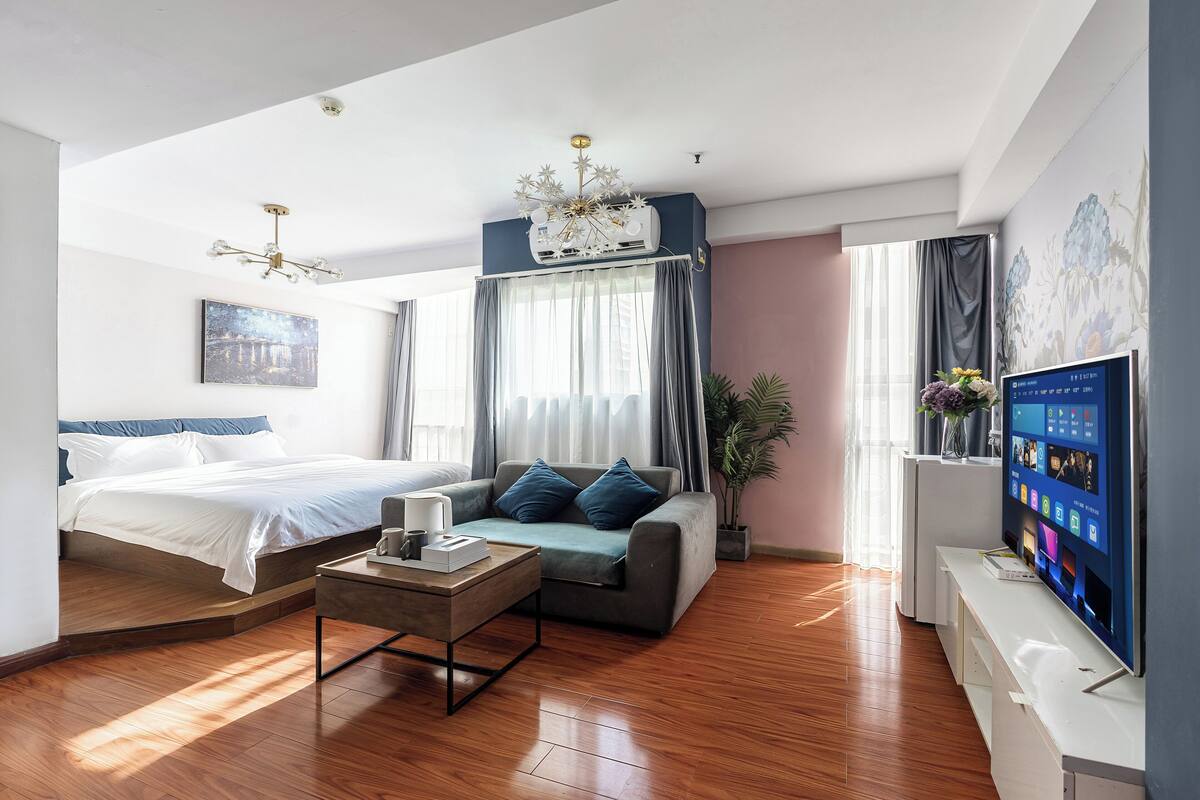 A modern hotel room with a bed, a sofa, a coffee table, a TV, and a large window with curtains.