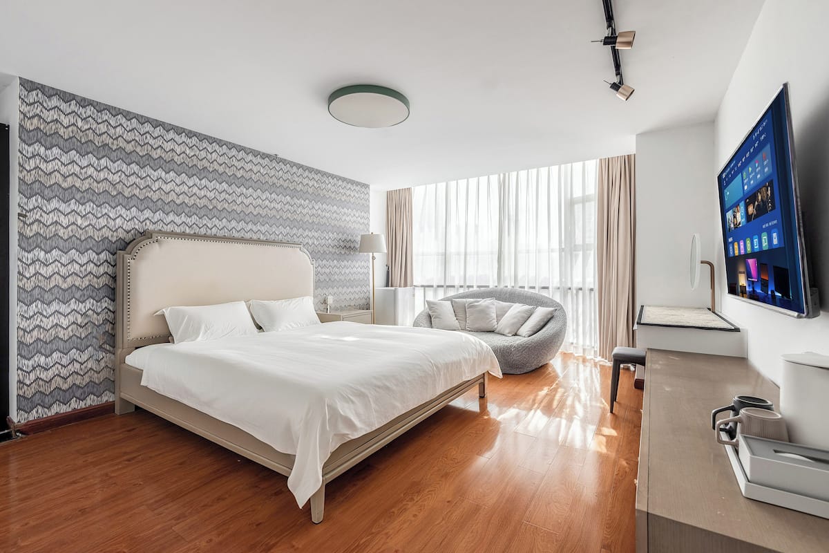 A modern hotel room with a large bed, a seating area, a TV, and a desk with a coffee machine.
