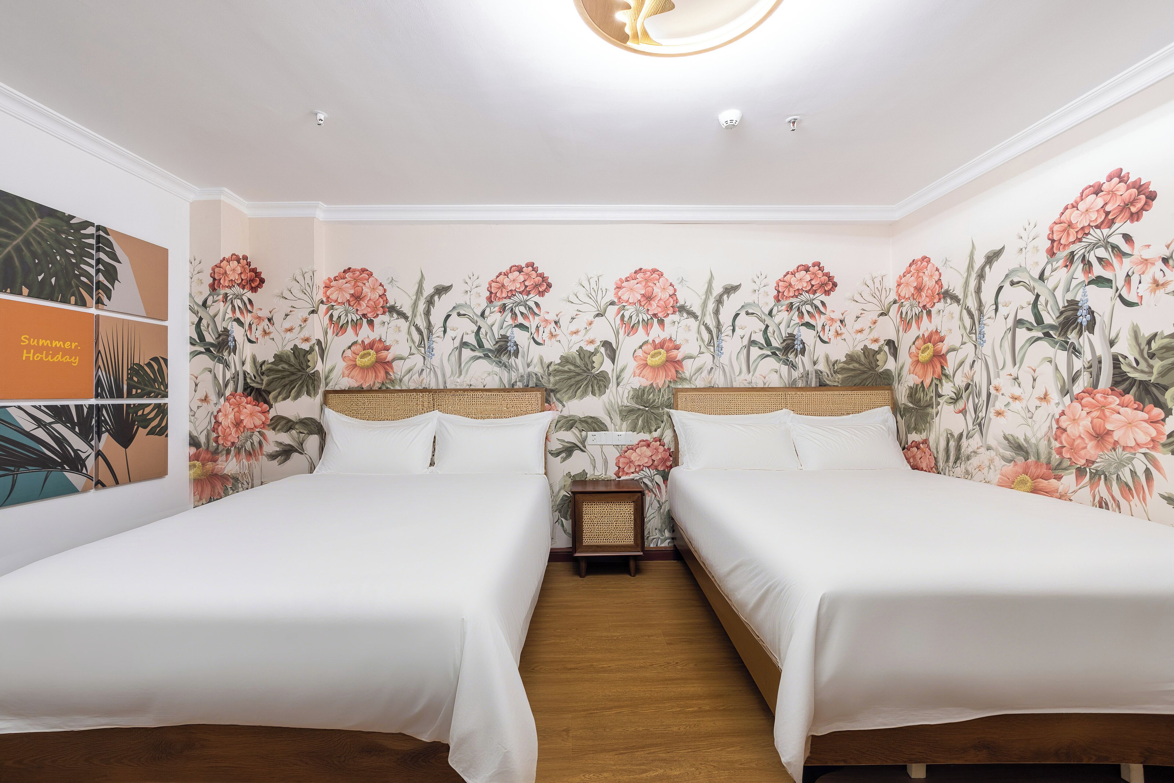 A room with two beds, floral wallpaper, a wooden floor, and a small table with a lamp.