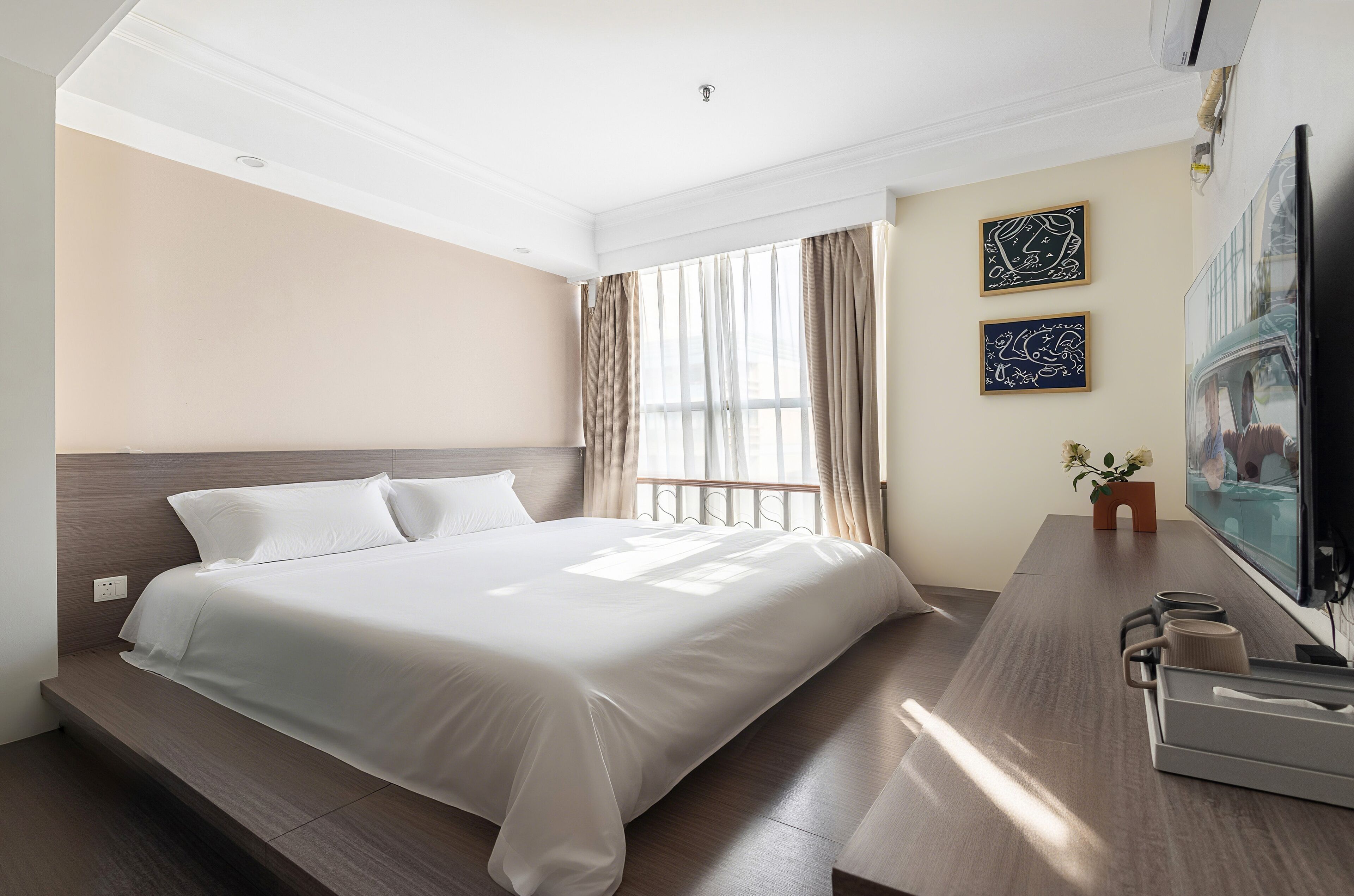 A hotel room with a large bed, a wooden headboard, a desk with a chair, a flat-screen TV, and two framed artworks on the wall.