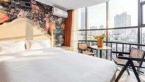 Habitación de hotel con una cama grande, una mesita con sillas, un balcón con vistas a la ciudad y un mural floral en la pared.