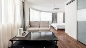 Una habitación de hotel moderna con una mesa de centro de madera oscura, un sofá beige, una cama y un ventanal con cortinas translúcidas.
