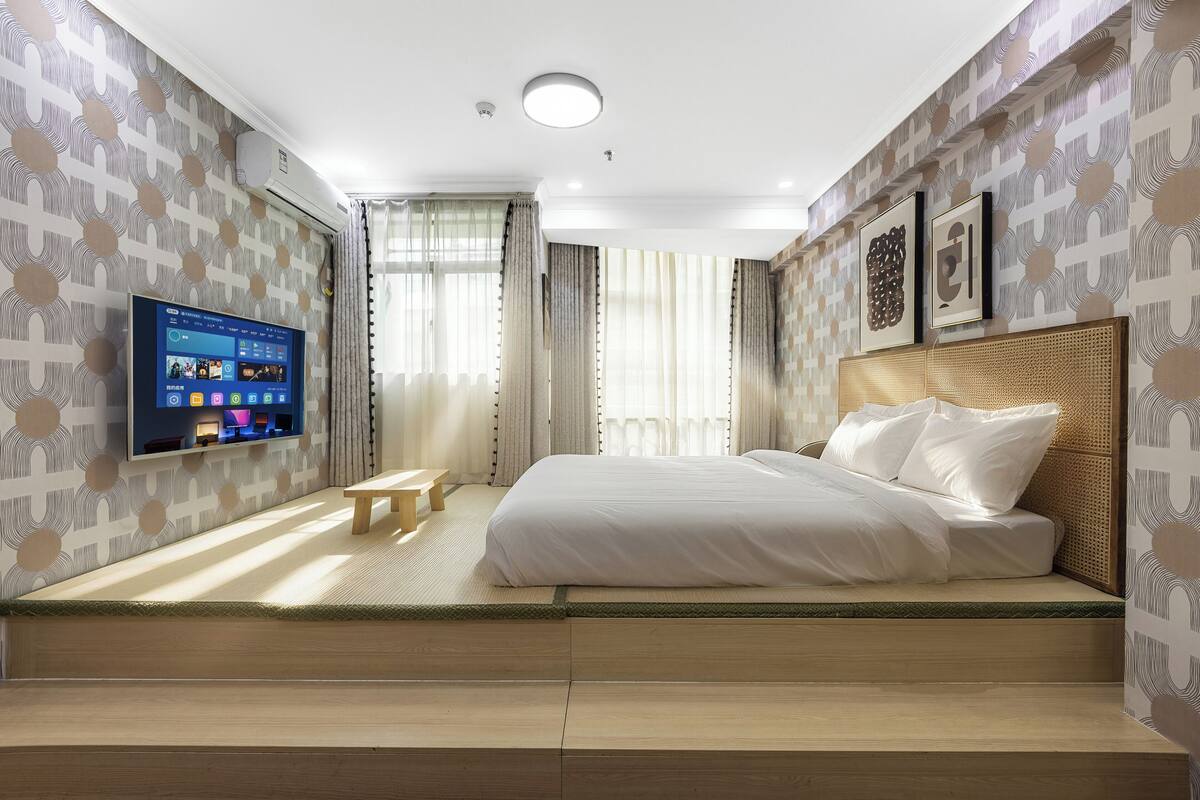 A modern hotel room with a bed, a TV, a small table, and a window with curtains.