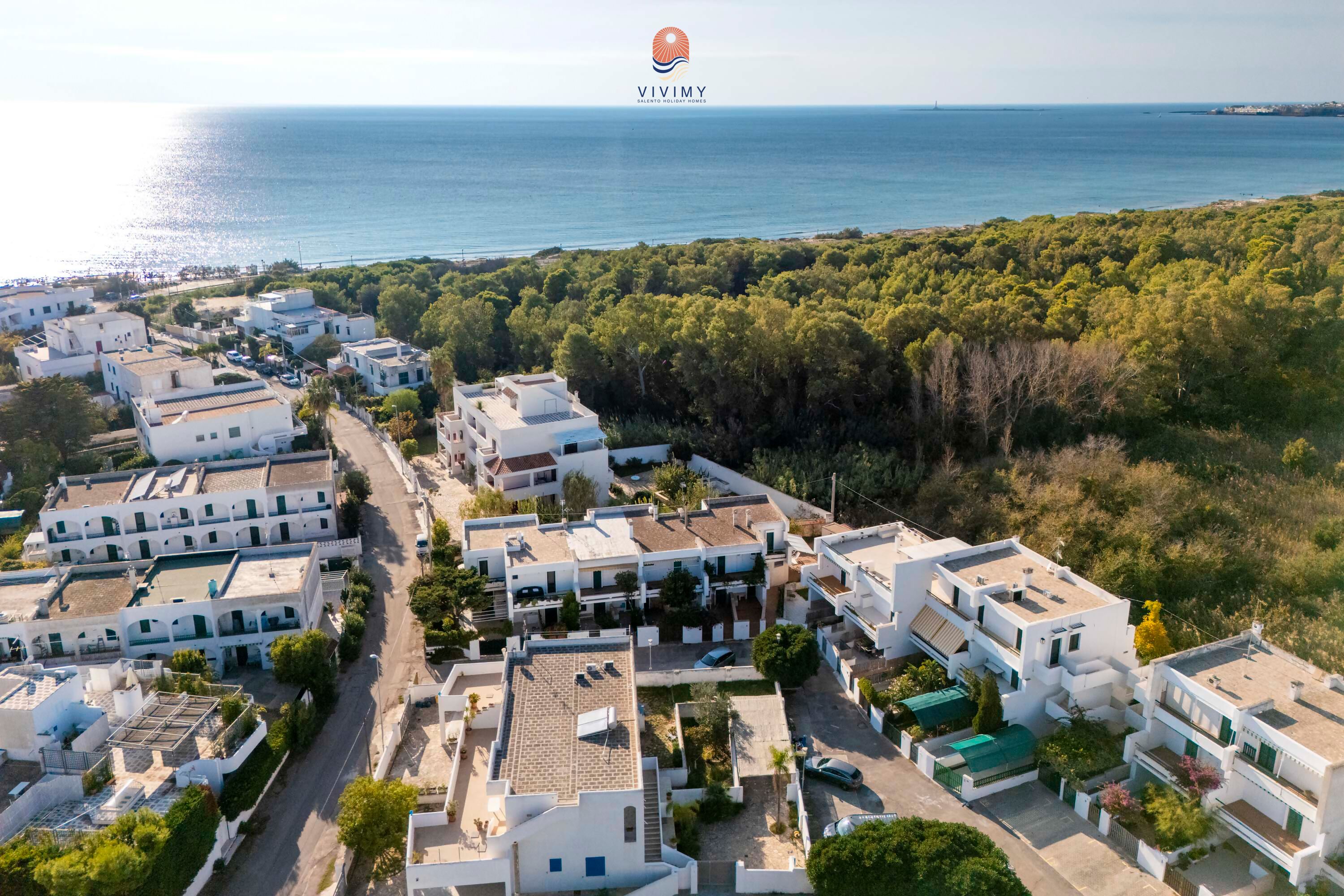 Vista aérea de una zona residencial costera con edificios blancos, una carretera y una zona boscosa al fondo.