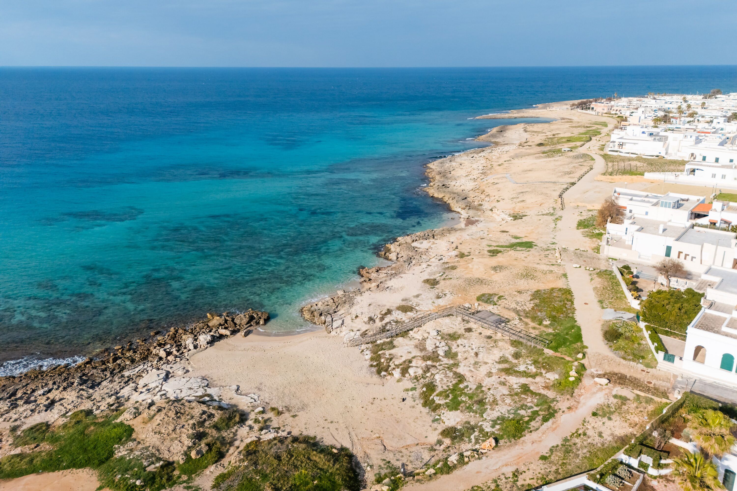 Vue aérienne d’une zone côtière avec des bâtiments blancs, une plage de sable et une eau d’un bleu limpide.