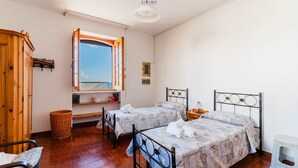 A bedroom with two beds, a wooden wardrobe, a window with an open shutter, a wicker basket, and a small stool.