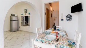 A dining area with a tiled table, white chairs, a TV mounted on the wall, and a kitchen with a refrigerator and microwave. - Nita House by Vivimy (Racale)