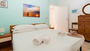 Un letto rifatto con cura, asciugamani arrotolati e un cappello, un comò in legno e un quadro raffigurante una barca a vela appeso al muro.