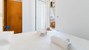 Ett hotellrum med en säng, två hoprullade handdukar, en trädörr, en blå lampa och utsikt över en balkong med möbler i rotting.