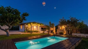 Ein Schwimmbad mit Holzterrasse, ein Haus mit Außenbeleuchtung und ein Baum im Vordergrund.