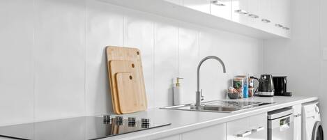 A modern kitchen with white cabinets, a stainless steel sink, a wooden cutting board, a coffee maker, and a washing machine.