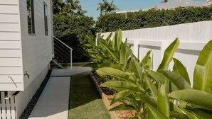 A white house with a paved walkway, a small garden with banana plants, and a white fence.