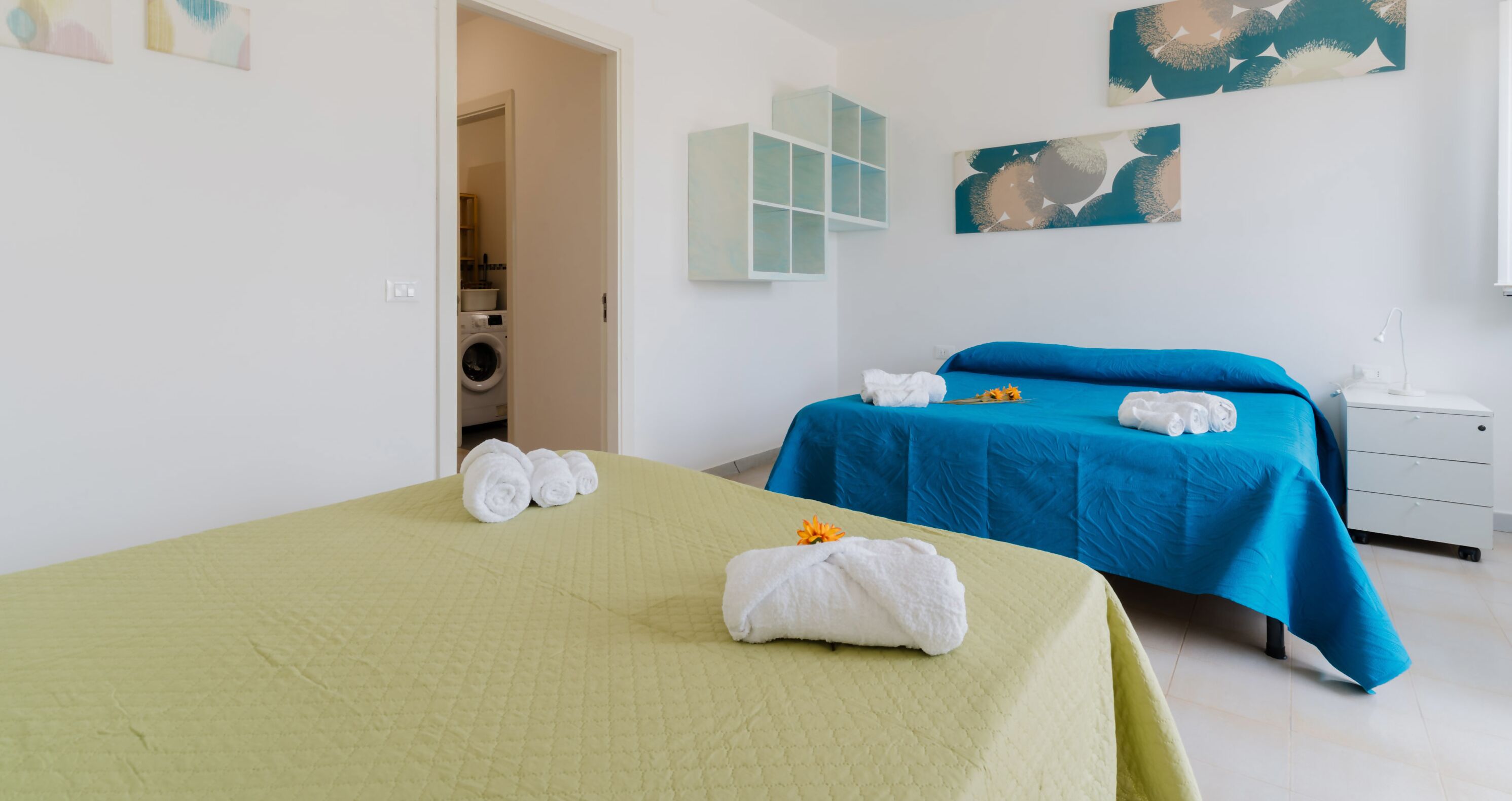 Ett hotellrum med två sängar, en tvättmaskin i hörnet och konstverk på väggarna.
