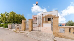 Ein verschlossenes Tor zu einem Anwesen mit einer Steinmauer, einem Metallgitter und einem gepflasterten Weg, der zu einem Gebäude mit einem Balkon führt.