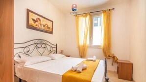 A neatly made bed with white linens and a yellow throw blanket, a wooden nightstand, a framed picture on the wall, and a window with yellow curtains. - Siren - Mediterranean Verandas (Torre Vado)