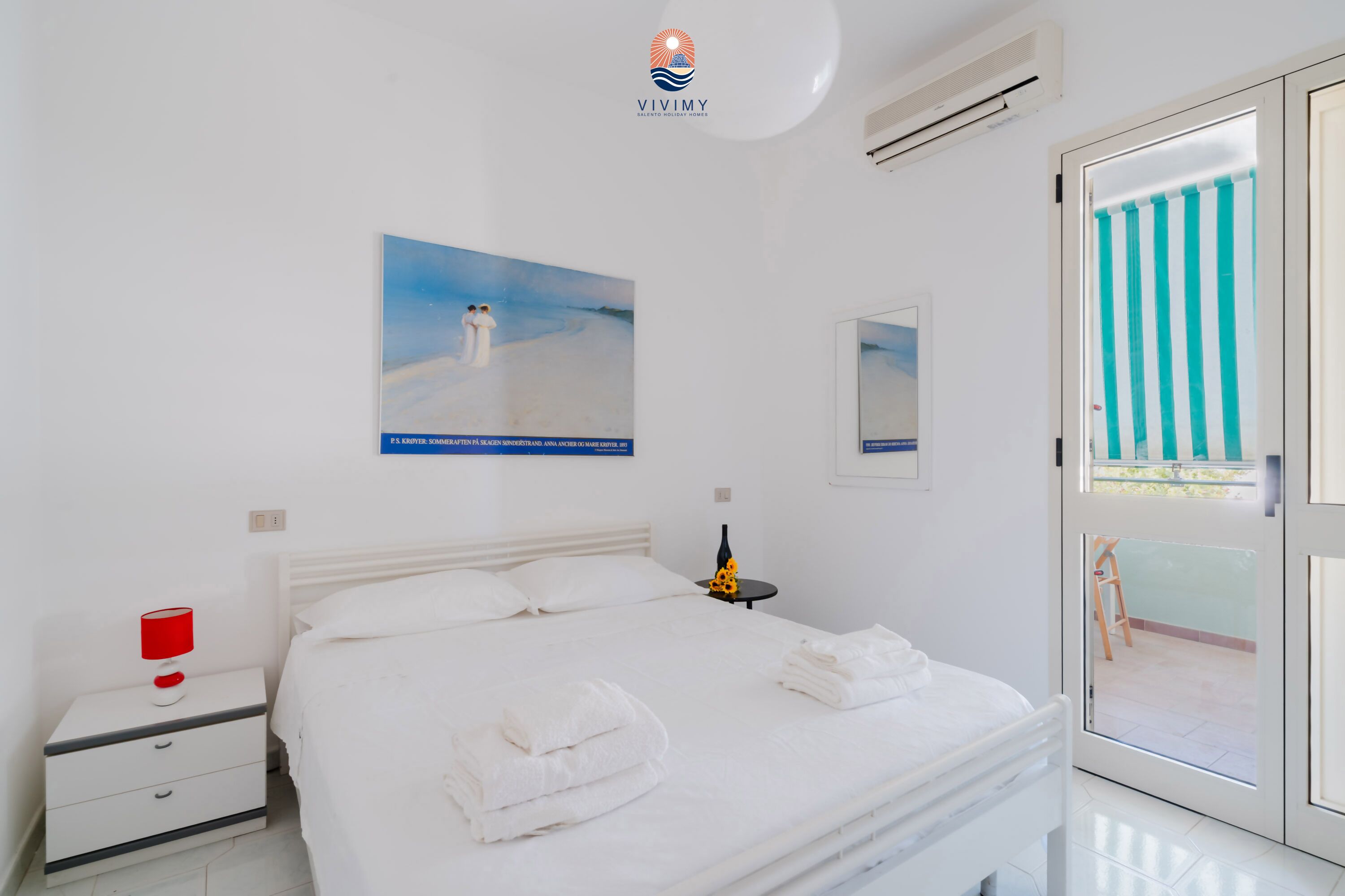 Ein Hotelzimmer mit Bett, Nachttisch, roter Lampe, Flasche und einem Bild von einer Strandlandschaft.