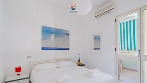 A hotel room with a bed, a nightstand, a red lamp, a bottle, and a painting of a beach scene. - Coral by Vivimy real estate (Gallipoli)