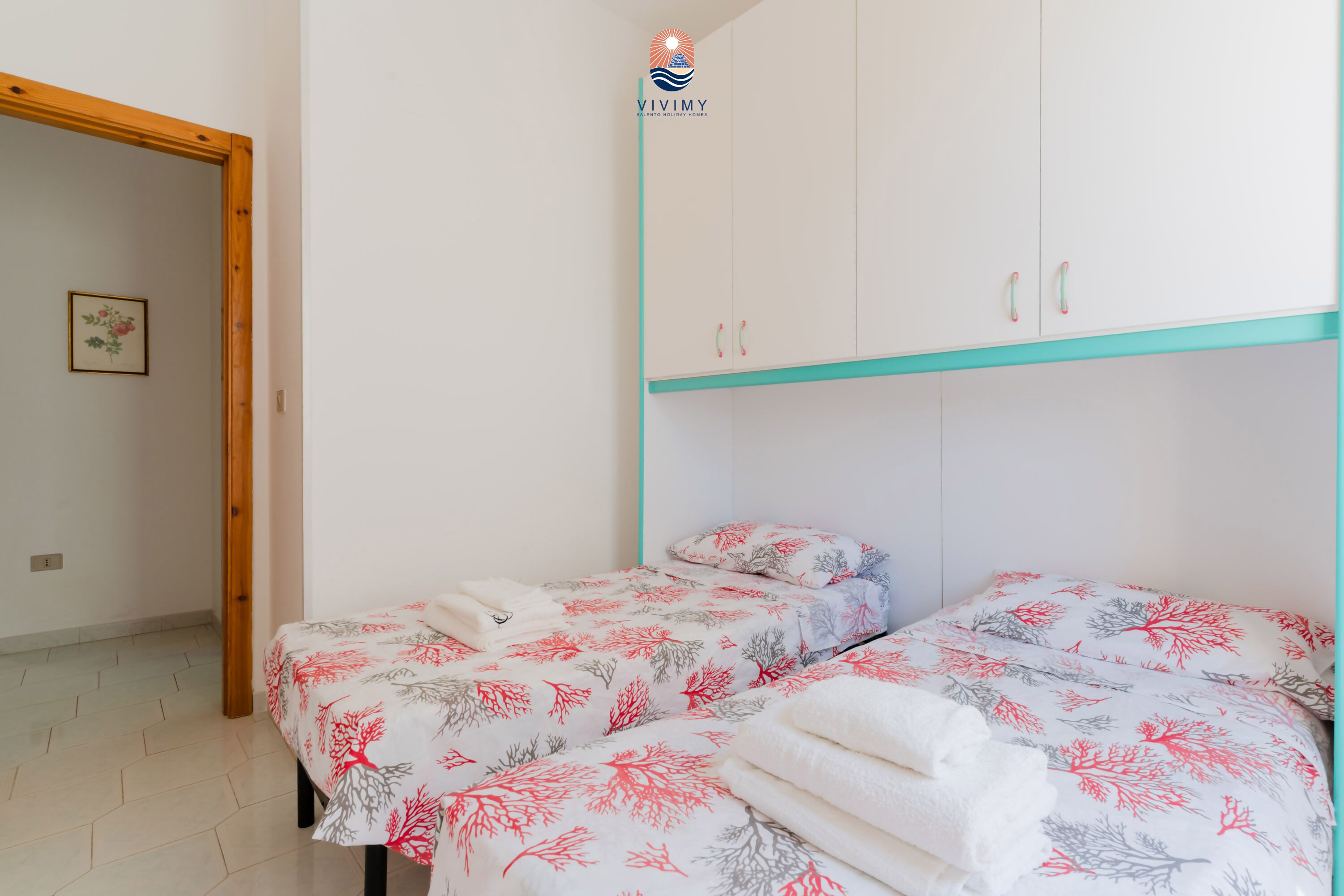 Une chambre avec deux lits, une porte en bois et un mur blanc orné d’un tableau encadré.