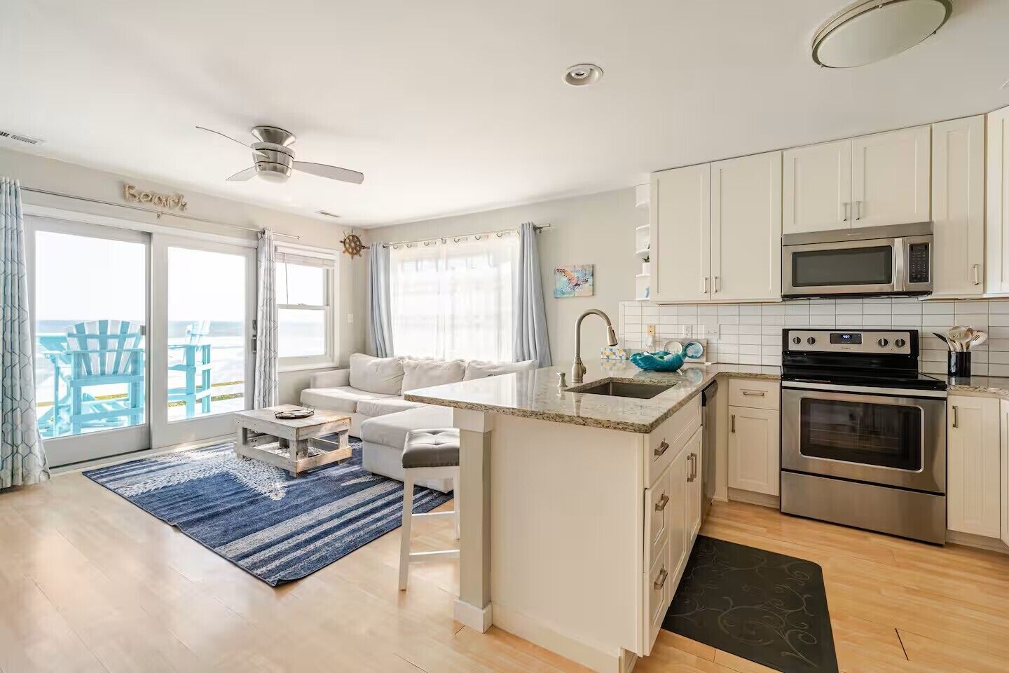 A modern kitchen with white cabinets, a stainless steel oven, and a microwave. There is a dining area with a white table and blue rug.