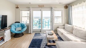 Ein Wohnzimmer mit Fernseher, blauem Sessel, Holztisch und einer Couch. Große Fenster mit Vorhängen bieten Blick auf den Strand.