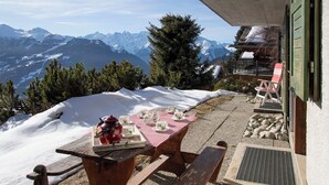 Una mesa de madera con tazas de té y una tetera sobre un mantel a cuadros, con vistas a montañas nevadas y un edificio de madera con una puerta verde.