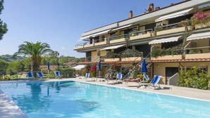 A swimming pool with lounge chairs and umbrellas, a multi-story building with balconies, and a clear sky. - Il Porto 19 - Mansarda sul Mare (Porto Azzurro)