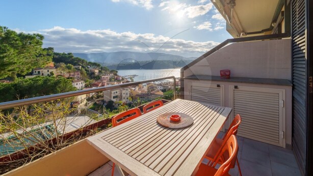 A balcony with a wooden table and orange chairs, overlooking a coastal town with buildings and a body of water. - Il Porto 19 - Mansarda sul Mare (Porto Azzurro)