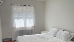 A bedroom with a wooden floor, a white bed, white nightstands, and a window with sheer curtains. - 2-bedroom apartment in cool Bronx with refreshing AC (Bronx)