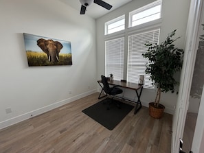 Una habitación amplia con piso de madera, un escritorio con silla, un ventanal con persianas, una planta en maceta y un cuadro de un elefante.