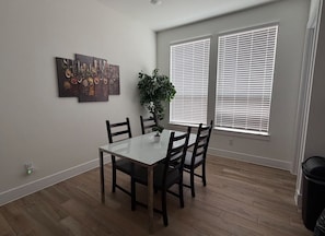 Un comedor con una mesa rectangular, cuatro sillas, una planta en maceta y una obra de arte en la pared.