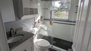 A compact bathroom with a white toilet, a granite countertop sink, a glass shower enclosure, and a window providing natural light. - The Guesthouse on Mills 50 (Orlando)