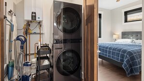 Een wasserette met gestapelde wasmachines en drogers, een plafondventilator, en een bed in de aangrenzende kamer met een blauw geruit sprei.