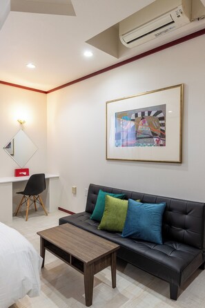 Ein modernes Hotelzimmer mit einer schwarzen Ledersofa, einem Holztisch, einem Schreibtisch mit Stuhl, einem gerahmten abstrakten Gemälde und einer Klimaanlage.