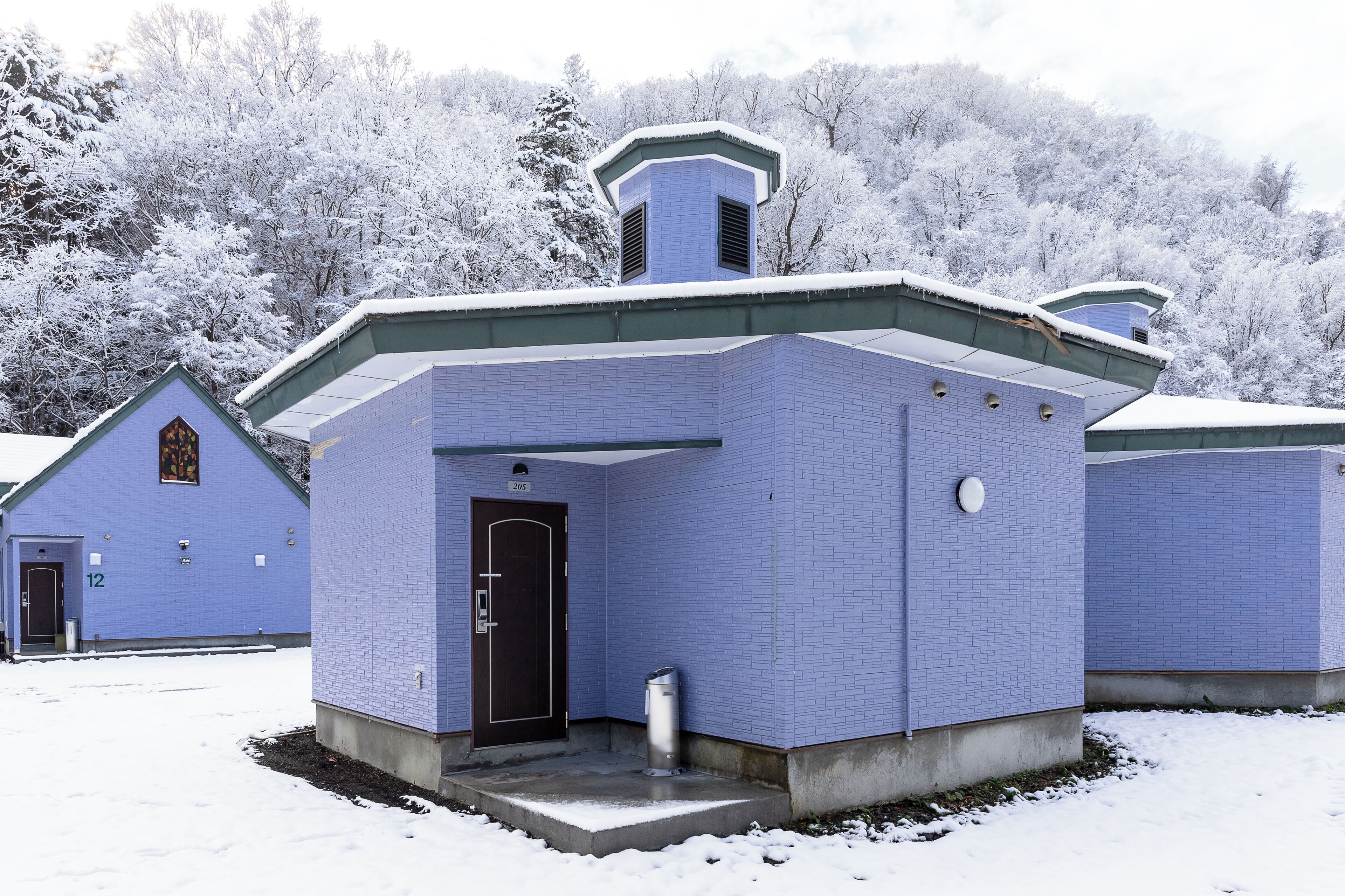 Un edificio azul con una puerta y una ventana pequeña, rodeado de árboles cubiertos de nieve.
