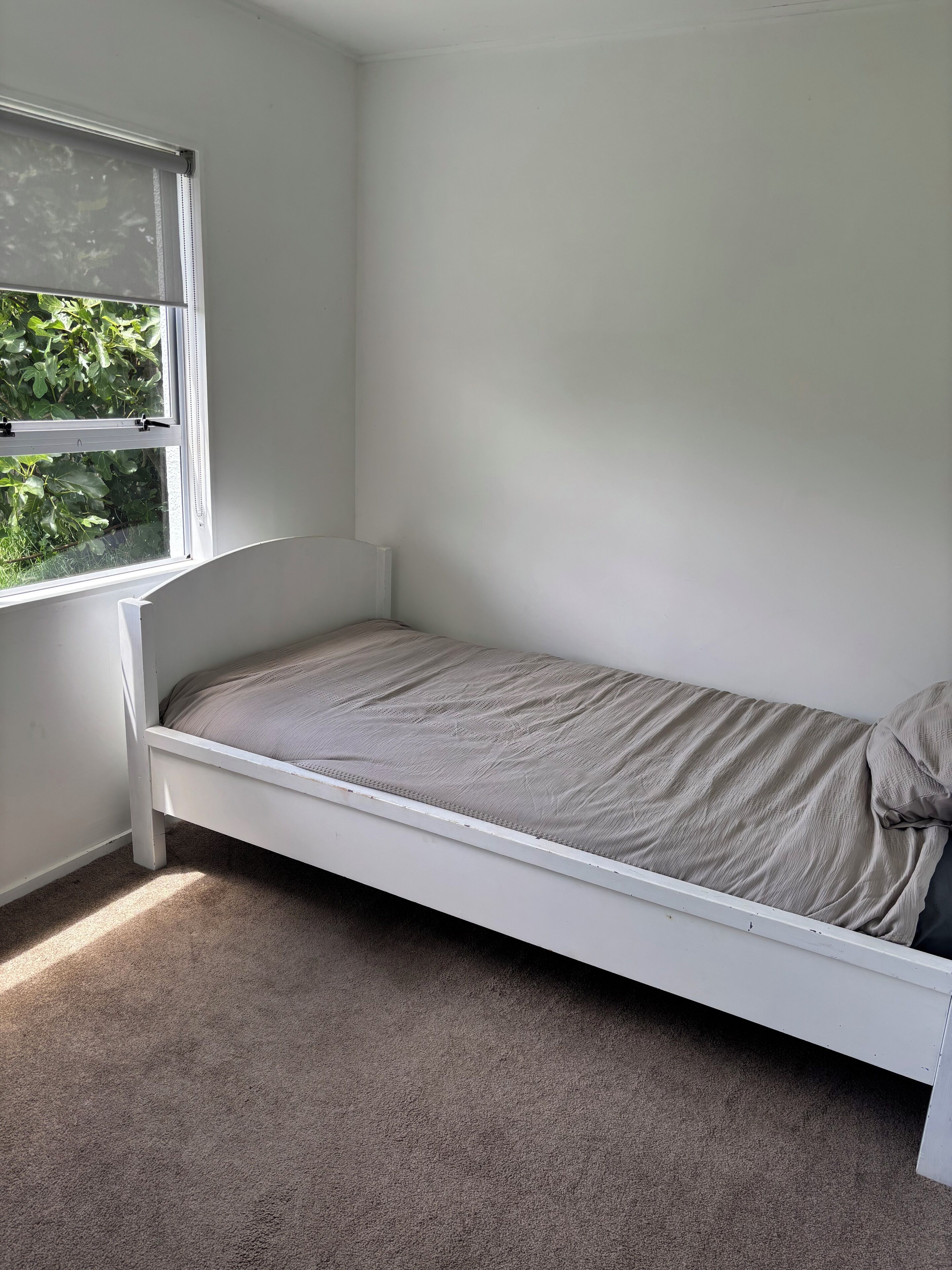 Een kleine kamer met een enkel bed, raam, vloerbedekking en witte wanden.