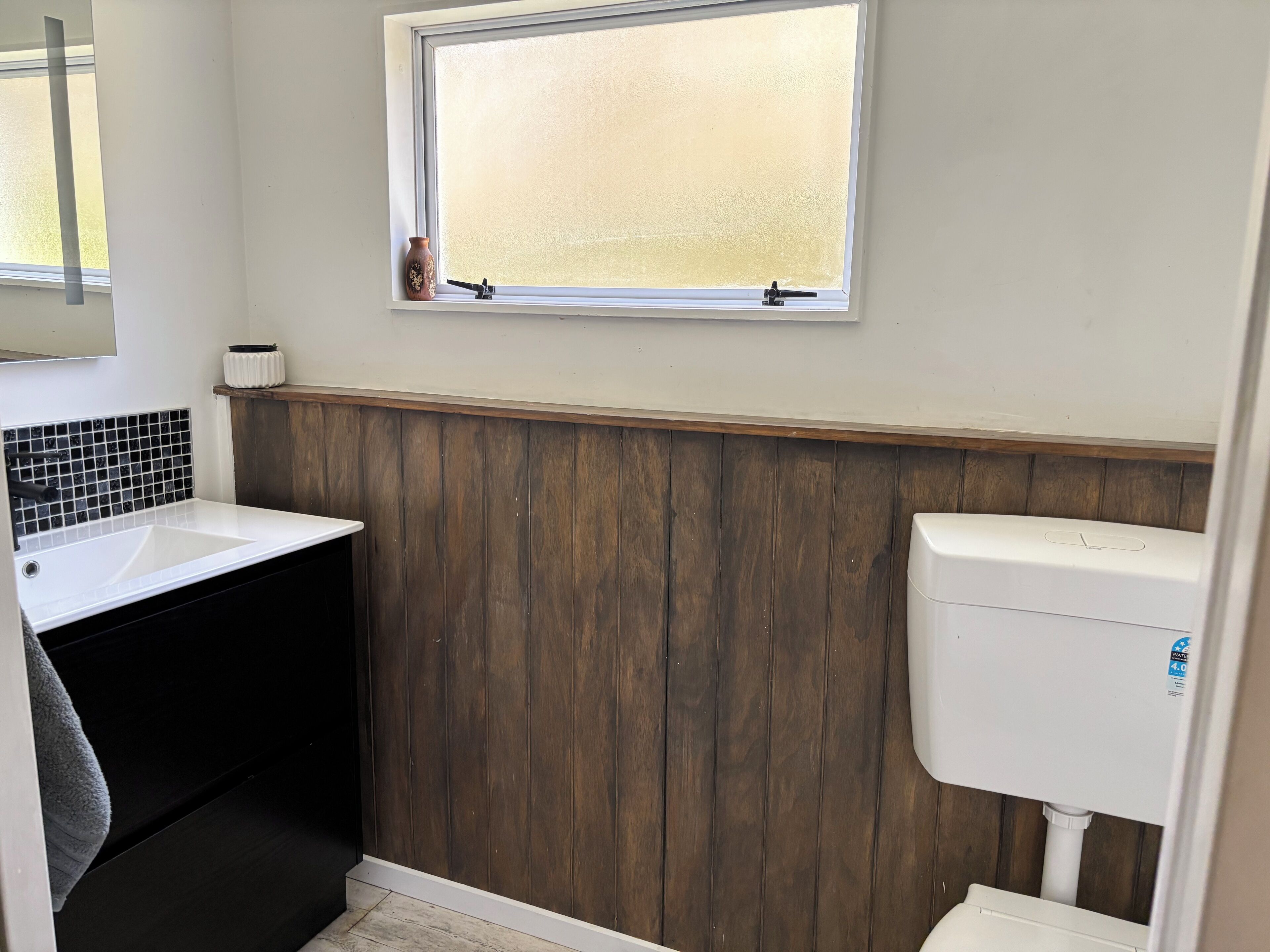 A bathroom with a white sink, wooden paneling, a toilet, and a window with blinds.
