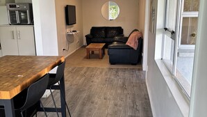 A modern kitchen with a built-in oven and microwave, a dining area with a wooden table and chairs, a living room with a sofa and a coffee table, and a large window with a view of the outdoors. - Spacious Three Bedroom Coastal Apartment in Army Bay (Whangaparāoa)