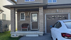 A two-story house with a garage, a front porch with a bench, and a white car parked in the driveway. - Cozy Queen Room Near Hospital | Quiet & Clean Home (Napanee)