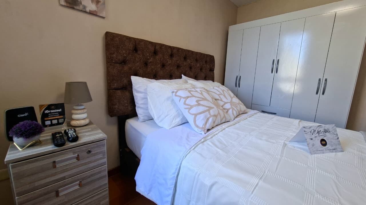 Camera da letto con testiera imbottita, biancheria bianca, comodino con lampada e armadio.