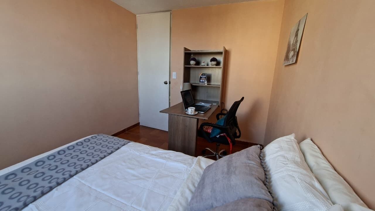 Um quarto pequeno com uma cama, uma escrivaninha com um laptop, uma cadeira e uma estante com objetos decorativos.