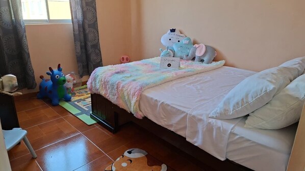 Un dormitorio con una cama, almohadas, una colcha de colores del arcoíris, peluches y una alfombra pequeña.