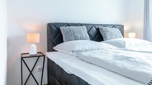 A neatly made bed with white linens and two grey pillows, a black upholstered headboard, and a small bedside table with a lamp. - Stylish 1BR 1LR Vienna Center Homes (Vienna)