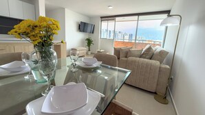 Un salon moderne avec une table à manger en verre, un canapé beige et une grande fenêtre donnant sur la ville.